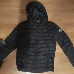 Pufferjacka från Vinson Polo Club - Säljer en svart pufferjacka från Vinson Polo Club med huva och dragkedja. Jackan har ett märke på ärmen och är perfekt för kyligare dagar. Den är quiltad och har en stilren design. Passar dig mellan 160-170cm