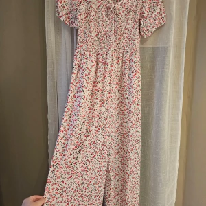 Blommig jumpsuit från FB Sister - Charmig blommig jumpsuit från FB Sister i vitt med röda och svarta blommor. Klänningen har korta ärmar och en smockad överdel med knytning framtill. Perfekt för en somrig look! 🌸 Oanvänd.