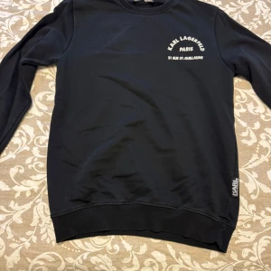 Svart tröja från Karl Lagerfeld - Snygg svart tröja från Karl Lagerfeld med tryck '21 RUE ST-GUILLAUME' både fram och bak. Tröjan har långa ärmar och en ribbad nederkant. Perfekt för en stilren look.