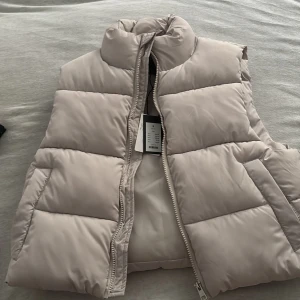 Beige dunväst med dragkedja - Snygg och stilren beige dunväst från lager 157. Västen har en praktisk dragkedja framtill och är perfekt för våren. Jackan är oanvänd med prislapp på. 