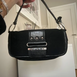 Svart axelväska från Guess - Svart axelväska från Guess med silverdetaljer och logotyp framtill. Väskan har en praktisk dragkedja och en stilren design som passar perfekt för olika tillfällen. Den är knappt använd och i nytt skick!