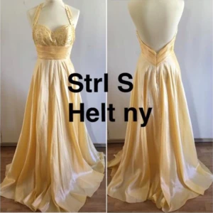 Gul balklänning med halterneck - Elegant gul långklänning med halterneck och öppen rygg. Klänningen har en vacker glittrig detalj på bysten och ett brett midjeband som framhäver figuren. Perfekt för en somrig bal eller fest. Använd 1 gång.