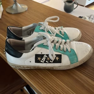 Vita och turkosa sneakers med stjärndetaljer - Snygga sneakers i vitt och turkost med svarta detaljer och stjärnor. Skorna har snörning och en cool 'Love' text på sidan. Perfekta för en avslappnad stil.