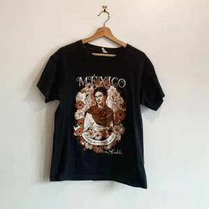 Svart t-shirt med Frida Kahlo-tryck - Cool svart t-shirt från King Tees med ett stort tryck av Frida Kahlo och texten 'Mexico'. Tillverkad i 100% bomull för en skön känsla. Perfekt för dig som vill ha en unik stil med konstnärlig touch.