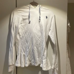 Vit hoodie från U.S. Polo Assn. - Säljer en stilren vit hoodie från U.S. Polo Assn. med en liten logga på bröstet. Tröjan har långa ärmar och en justerbar huva med randiga snören. Tillverkad i 100% bomull för en bekväm känsla. Perfekt för en avslappnad stil.