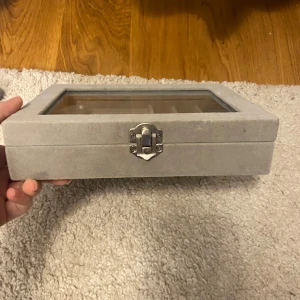 Grå smyckeskrin med glaslock - Elegant grå smyckeskrin med glaslock och lås. Inuti finns det plats för ringar och flera fack för andra smycken. Perfekt för att organisera och visa upp dina favoritsmycken.