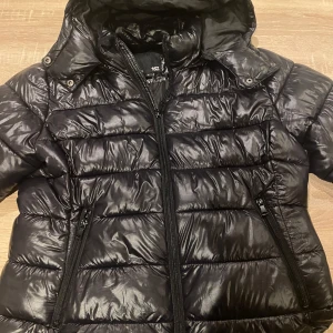 Svart pufferjacka från 157 - Snygg svart pufferjacka från 157 med dragkedja och huva. Jackan har en glansig finish och två praktiska fickor med dragkedjor. Perfekt för kyliga dagar när du vill hålla stilen.