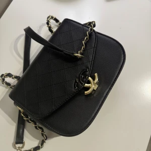 ”INTE DEN RIKTIGA ”Svart quiltad axelväska med kedjedetaljer - Snygg svart axelväska med quiltat mönster och guldfärgade kedjedetaljer. Väskan har en elegant design med en praktisk ficka på baksidan och ett rött innerfoder. Perfekt för att ge din outfit en lyxig touch.