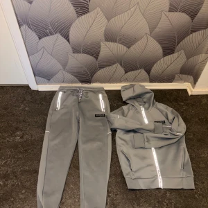 Grå Tracksuit från McKenzie - Säljer en skit snygg mckenzie tracksuit med färgen baby blue. Plaggen har reflex som syns när flash kommer på och med ljus i mörkret. Linjen vid koftans zip linje är lite sliten men finns ändå lite kvar. Nypris runt 1600kr, köpt i London. Unik set som inte finns längre. Byxorna är storlek 12/13 years old och koftan 10/12 years old. Tveka inte att höra av dig vid intresse.