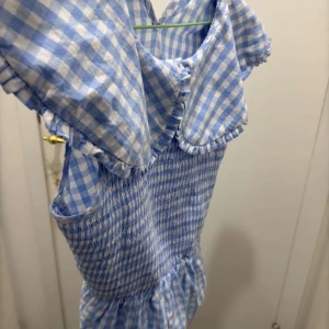 Blå och vit rutig blus - Charmig blå och vit rutig blus  med korta ärmar och en söt krage. Blusen  har en smockad midja som ger en fin passform och volangdetaljer vid kragen. Perfekt för en somrig look!