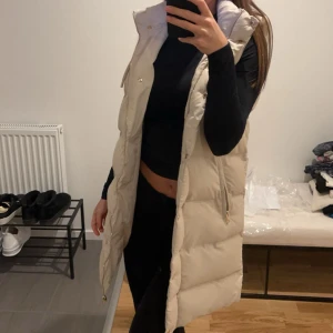 Beige dunväst med huva - Säljer en snygg beige dunväst med huva och dragkedja. Västen har en lång passform och knappar framtill. Perfekt för kyligare dagar när du vill hålla stilen. Från zara