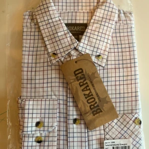 Rutig tattersall skjorta från Brokared ny i förpackning  - Snygg rutig tattersall skjorta från Brokared i vitt med blå och röda rutor. Ny i förpackningen. Skjortan har långa ärmar och knappar framtill samt på bröstfickan. Perfekt för en stilren look.