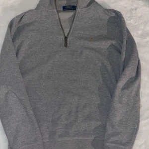 Grå tröja med dragkedja från Ralph Lauren - Säljer en stilren grå tröja från Ralph Lauren med halv dragkedja och broderad logga på bröstet. Perfekt för en avslappnad men snygg look. Tröjan har långa ärmar och är tillverkad i mjukt material.