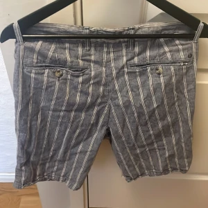 Randiga shorts från Dressmann - Snygga randiga shorts från Dressmann i en skön linneblandning. Perfekta för varma dagar med sin luftiga passform och stilrena design. Shortsen har knappar och dragkedja framtill.