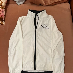 Vit vindjacka med tryck - Snygg vit vindjacka med svart dragkedja och tryck på bröstet och ryggen. Jackan har en hög krage och långa ärmar, perfekt för blåsiga dagar. Den har en stilren design med nautiskt tema.