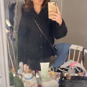 Svart kappa med knappar - Säljer en stilren svart kappa med knappar framtill. Jackan har en klassisk design med långa ärmar och en elegant passform. Perfekt för kyligare dagar och ger en sofistikerad look.