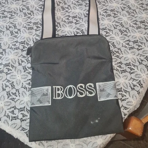 Svart axelväska från Boss - Snygg svart axelväska från Boss med vita detaljer och logotyp framtill. Väskan. Säljer den då den inte används' en av bokstäverna e lösa men går bara att limma fast lite!