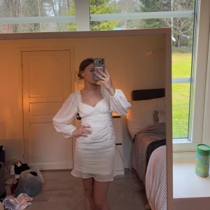 Vit klänning med puffärmar - Säljer en stilren vit klänning med långa puffärmar och v-ringad design. Klänningen har en kort längd och en figurnära passform som framhäver formen . Perfekt för studenten! 