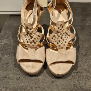 Beige och guldiga klackskor med nitar - Eleganta klackskor i beige mocka med glittriga och guldfärgade detaljer. Skorna har en spetsig tå och dekorativa nitar på ovansidan. Perfekta för en festlig kväll.