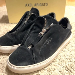 Svarta sneakers från Axel Arigato - Snygga svarta sneakers från Axel Arigato i mocka med dragkedja framtill. Skorna har en vit sula och är handgjorda i Portugal. Perfekta för en stilren look.