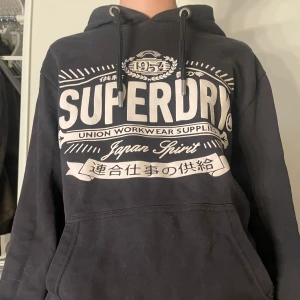 Svart hoodie från Superdry - Säljer en svart hoodie från Superdry med stor vit logga och text på framsidan. Tröjan har en klassisk känguruficka och justerbar huva med dragsko. Perfekt för en avslappnad stil. NYPRIS 1000 vårt 400