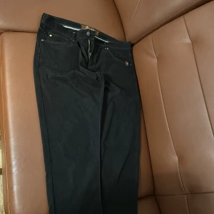 Svarta byxor från Sand - Snygga svarta byxor från Sand med superstretch för en bekväm passform. Byxorna har en slim fit och är utrustade med dragkedja och knappar. Perfekta för en stilren look.