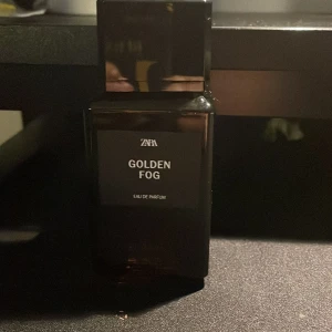 Golden Fog Eau de Parfum från Zara - Elegant parfymflaska i mörkt glas med namnet Golden Fog. Flaskan har en stilren design med en avtagbar fyrkantig kork. Perfekt för den som söker en sofistikerad doftupplevelse.