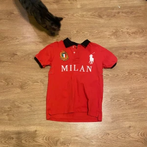 Ralph Lauren ”MILAN” Polo  - Snygg röd pikétröja från Ralph Lauren med svart krage och MILAN-tryck i vitt. Tröjan har korta ärmar och en broderad logga på bröstet. Perfekt för en sportig look! Pris kan diskuteras vid snabb affär