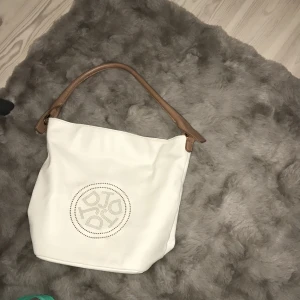 Vit axelväska med bruna detaljer - Snygg vit axelväska med bruna handtag och detaljer. Väskan har en praktisk dragkedja på baksidan och ett blommigt mönster inuti. På den sista bilden syns några få små stretck, vet ej vad de kommer ifrån. Men de syns inte jättemycket. 