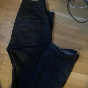 Svarta Nudie jeansbyxor - Snygga svarta Nudie jeansbyxor med en klassisk femficksdesign. De har en normal passform och är perfekta för en stilren look. Byxorna har en dragkedja och knapp framtill.