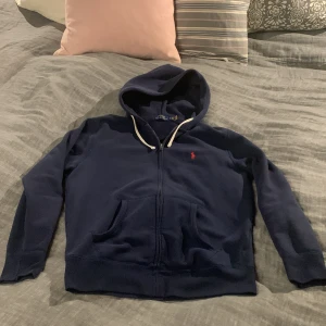 Mörkblå hoodie från Ralph Lauren - Säljer en snygg mörkblå hoodie från Ralph Lauren med dragkedja och klassisk logga på bröstet. Tröjan har långa ärmar och en praktisk huva med snören. Den är i skick 8/10 skulle jag säga. Nypris ligger på lite över 2000 men säljer för 550 kr