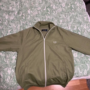 Olivgrön track jacket från Fred Perry - Säljer min Fred Perry tröja då den inte passar mig längre. Den är storlek S och knappt använd, inga fläckar. Priset är inte fast, tar emot bud!