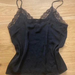 Svart linne med spetsdetaljer - Snyggt svart linne med smala axelband och spetsdetaljer längs halsringningen. Perfekt för en stilren look.