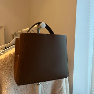 Brun handväska från Gina Tricot - Snygg brun handväska från Gina Tricot i syntetmaterial. Väskan har en stilren design med en praktisk dragkedja inuti och en knappstängning. Perfekt för att bära dina dagliga nödvändigheter med stil. Långa band medföljer!❤️