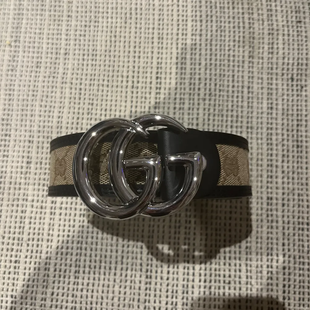 Snyggt bälte från Gucci med en iögonfallande silverfärgad GG-spänne. Bältet har ett klassiskt mönster i beige och svart, perfekt för att lyfta vilken outfit som helst. Tillverkat i skinn med detaljerad design.. Asusteet.