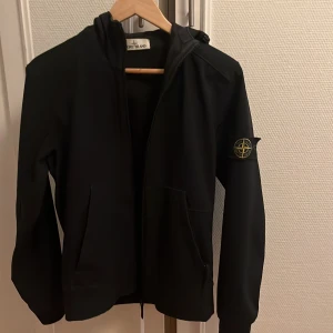 Svart jacka från Stone Island - Säljer en stilren svart jacka från Stone Island med dragkedja och huva. Jackan har det ikoniska märket på ärmen och är perfekt för en modern look. Passar bra till både vardag och mer avslappnade tillfällen. Storlek xs/s