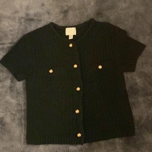 Elegant kort stickad kofta  - Snygg svart stickad kofta från H&M med korta ärmar och dekorativa guldknappar. Perfekt för både kalas, middagar eller vardag. Man får även med en extra guldknapp utifall en knapp skulle ramla av. Använd 2 gånger. Säljer för att jag har tröttnat. 