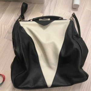 Svart och beige väska från Norrback - Snygg svart och beige väska från Norrback med praktisk dragkedja. Väskan har en unik design med kontrasterande färger och en justerbar axelrem. Perfekt för att bära dina dagliga nödvändigheter med stil.