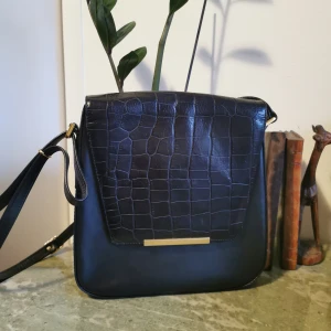 Svart axelväska med krokodilmönster - Snygg svart axelväska med krokodilmönstrad framsida och gulddetaljer. Uppdaterar med mått, men skriv gärna om jag inte hunnit. Lite tillknycklad i rämmen pga har legat i en flyttkartong. 💼