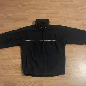 Svart jacka från Blend - Säljer en svart jacka från Blend med dragkedja och hög krage. Jackan har en enkel design med en vit linje som detalj på framsidan och baksidan. Perfekt för kyligare dagar.