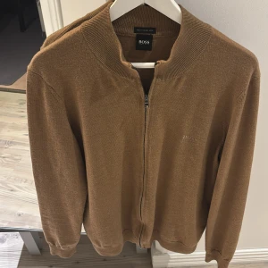 Brun kofta från Hugo Boss - Snygg brun kofta från Hugo Boss i finaste italienska garnet. Den har en dragkedja framtill och ribbade muddar vid ärmslut och nederkant. Perfekt för en stilren look och bekvämlighet vardag i topp skick.