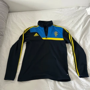 Svart och blå långärmad tröja från Adidas - Säljer en svart och blå långärmad tröja från Adidas med Sveriges fotbollsförbunds logga på bröstet. Tröjan har gula detaljer och en halv dragkedja framtill. Perfekt för fotbollsfans! 💙💛. Står ingen storlek på tröjan men skulle säga att den är s/m är 173 och den e ytterst lite stor på mig