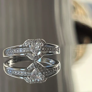 Äkta Silverring med hjärtformad sten - Elegant silverring med en stor hjärtformad sten i mitten och små gnistrande stenar längs bandet. Perfekt för speciella tillfällen eller som en kärleksfull gåva. Ringen har en modern och stilren design som fångar ljuset vackert. Äkta silver 925