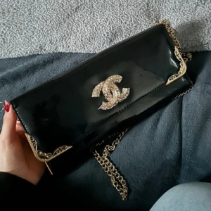 Svart glansig kuvertväska med kedja - Elegant svart kuvertväska i glansigt material med gulddetaljer och en kedja för axelrem. Väskan har ett stort emblem på framsidan och flera fack inuti för praktisk förvaring.