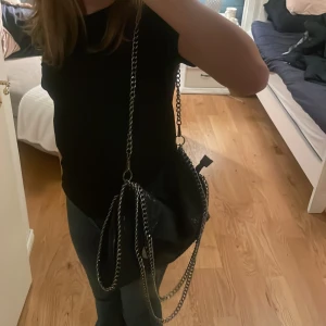 En liknade Stella McCartney! - Snygg svart axelväska med kedjedetaljer och dragkedja. Väskan har en glittrig yta och en praktisk storlek för vardagsbruk. Perfekt för att ge din outfit en edgy touch.