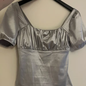 Silverfärgad topp - Snygg silverfärgad topp. Den har en elegant fyrkantig urringning och en glansig finish som passar perfekt för festliga tillfällen. Toppen är i bra skick och ger en lyxig känsla.