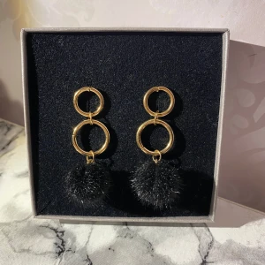 Guldörhängen med svarta bollar - Snygga guldörhängen med dubbla ringar och fluffiga svarta bollar. Perfekta för att ge en snygg touch till din outfit. Lätta att bära och passar till många olika stilar.