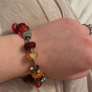 Färgglatt armband med pärlor från Pandora  - Snyggt armband med utbytbara pärlor i rött, orange och gult. Armbandet har en silverfärgad kedja och flera dekorativa pärlor som ger en unik look. Perfekt för att lägga till en färgklick till din outfit.dra inte för hårt eftersom pärlorna kan lossna, man måste sätta fast armbandet mycket hårt.