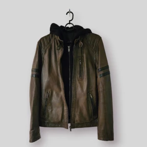 Brun skinnjacka från Saki - Snygg brun skinnjacka från Saki med svarta detaljer och dragkedjor. Jackan har en avtagbar huva och "hoodie". Perfekt för en cool och avslappnad stil. Helt ny!! Kostade över 2000 ny.