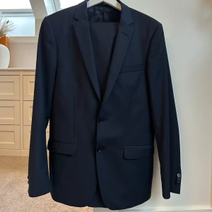 Marinblå kostym - Snygg mörkblå kavaj i klassisk stil med två knappar framtill. Perfekt för både formella och semi-formella tillfällen. Kavajen har en stilren design med fickor och är i mycket bra skick. Passar utmärkt till höst och vår.
